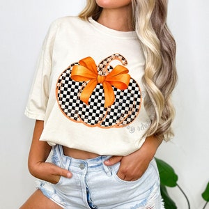 May include: Cream-colored t-shirt featuring a pumpkin design. The pumpkin has a black and white checkered pattern with an orange bow and stem. The design is centered on the shirt, creating a festive fall look.