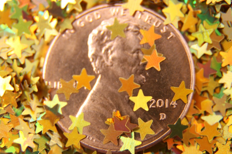 May include: A copper-colored US penny with the words "In God We Trust" and "2014 D" inscribed on it. The penny is surrounded by gold glitter stars.