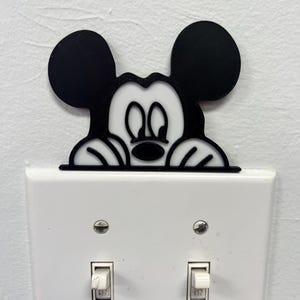 May include: A black and white Mickey Mouse light switch cover. The cover is designed to fit over a standard light switch and features the iconic character's face and ears. The light switch cover is a fun and decorative way to add a touch of Disney to any room.