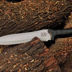 May include: A black-handled knife with a silver blade and a serrated edge. The blade has a logo engraved on it. The knife is lying on a piece of wood with wood shavings around it.