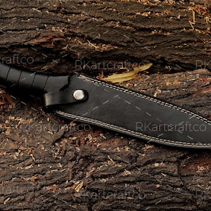 May include: A black leather knife sheath with white stitching detail. The sheath is lying on a piece of rough-hewn wood.
