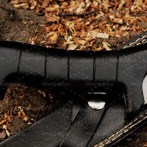 May include: A black leather sheath with white stitching detail. The sheath is partially open, revealing a black handle with a textured grip. The handle is attached to a silver metal buckle.