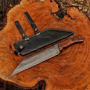 May include: A long, sharp, silver knife with a wooden handle and a black leather sheath. The knife is lying on a wooden surface.