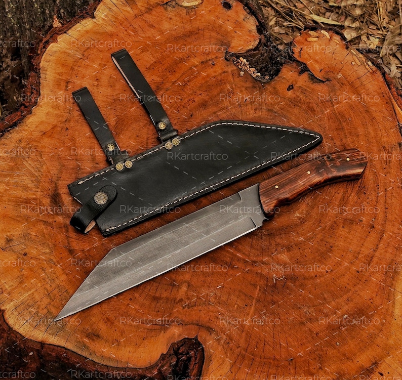 May include: A long, sharp, silver knife with a wooden handle and a black leather sheath. The knife is lying on a wooden surface.