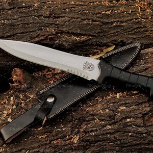 May include: A knife with a black handle and a silver blade, featuring a black leather sheath. The blade has a serrated edge and a decorative design near the handle.