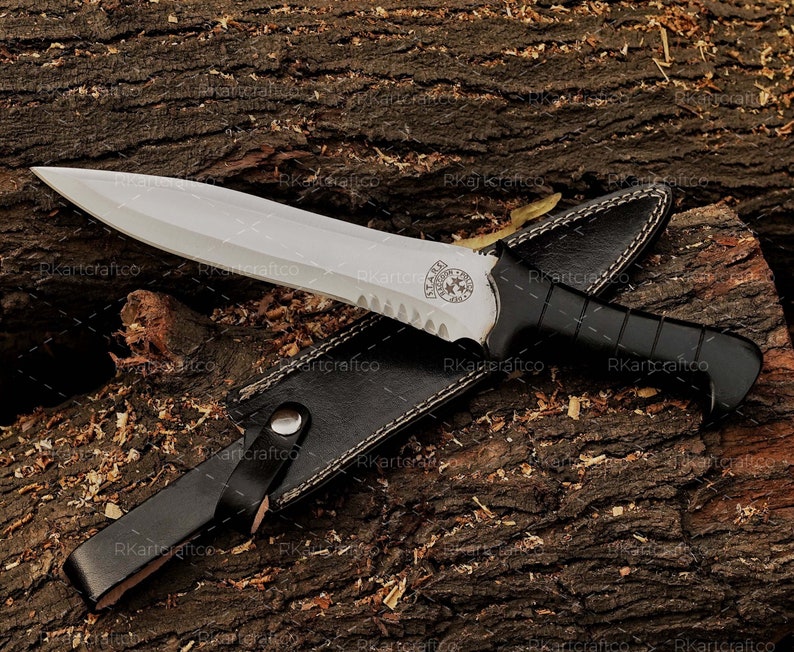 May include: A knife with a black handle and a silver blade, featuring a black leather sheath. The blade has a serrated edge and a decorative design near the handle.