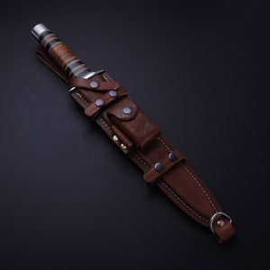 May include: A brown leather sheath with a knife inside. The sheath has multiple straps and snaps. The knife has a wooden handle with a black and silver blade.
