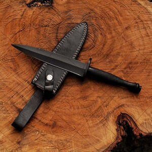 May include: A black dagger with a black handle and a leather sheath. The dagger is lying on a wooden surface. Close-up of custom D2 steel blade with sharp spear-point edge