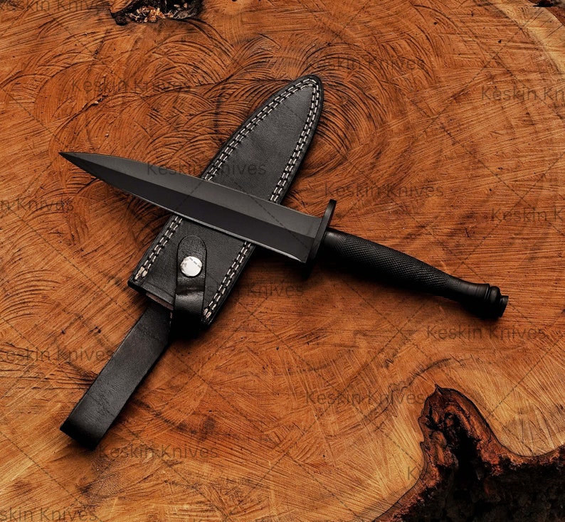 May include: A black dagger with a black handle and a leather sheath. The dagger is lying on a wooden surface. Close-up of custom D2 steel blade with sharp spear-point edge