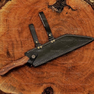 May include: A brown wooden-handled knife with a black leather sheath. The sheath has a strap with brass accents and stitching. The knife is lying on a wooden surface.