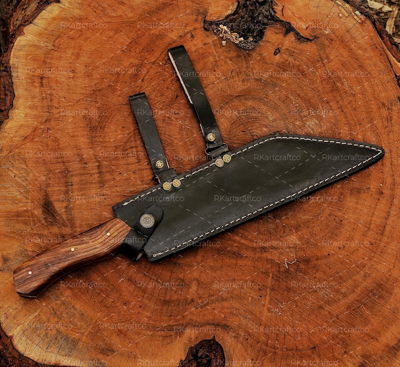 May include: A brown wooden-handled knife with a black leather sheath. The sheath has a strap with brass accents and stitching. The knife is lying on a wooden surface.