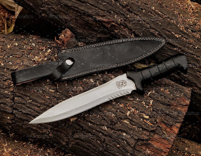 May include: A silver knife with a black handle and a serrated blade. The knife has a skull and crossbones logo on the blade. The knife is in a black leather sheath.