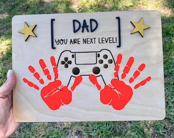Puede incluir: Un letrero de madera con el texto "[DAD] You are next level!" en letras negras. El letrero también presenta una ilustración en blanco y negro de un mando de videojuego y dos huellas de manos rojas.
