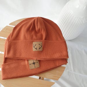 Könnte beinhalten: Zwei rostfarbene Mützen mit gerippter Struktur liegen auf einer Holzoberfläche. Jede Mütze hat ein quadratisches, beiges Patch. Eines zeigt ein Hirschgeweih-Design, das andere eine Windrose. Die Mützen haben einen warmen, erdigen Farbton.