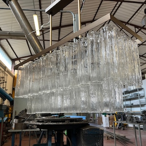 Puede incluir: Una gran lámpara de araña moderna con un marco rectangular de metal y muchos tubos de vidrio transparente que cuelgan de ella. La lámpara de araña está en un taller.