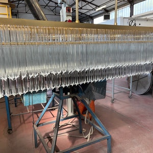 May include: A large, rectangular chandelier with a gold metal frame and hundreds of clear glass crystals hanging from it. The chandelier is suspended from the ceiling and is being assembled in a workshop.