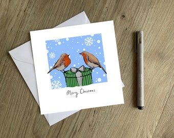May include: A Christmas card featuring two robins perched on a green and white striped gift box. The background is a light blue with white snowflakes. The card reads "Merry Christmas".