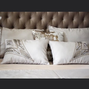 May include: A set of four decorative throw pillows with a light beige fabric and intricate silver and gold embroidery. The pillows are arranged on a tufted headboard with a light brown fabric.