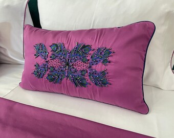 May include: A pink pillow with a decorative embroidered design featuring peacock feathers in shades of green, blue, and purple. The pillow has a navy blue trim.