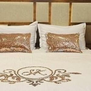 May include: White bed with a white duvet cover and two white pillowcases. The duvet cover has a gold embroidered design with the initials "SK". Two gold sequin pillows are on the bed.
