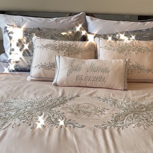May include: A pink bed with a white and silver embroidered design. The bed is made up with white and silver pillows, including one that says "Just Married 05.01.2024".