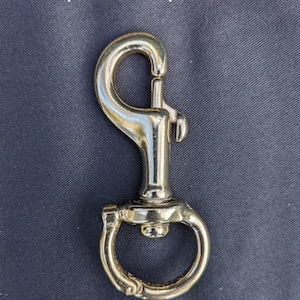 May include: A shiny gold-colored snap hook with a spring-loaded clasp and a circular base. The text "Replacement Snaps Available" is at the top. The hook is designed for attaching to various items.
