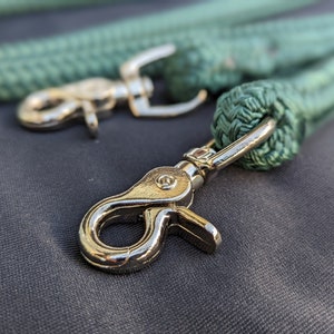 May include: Close-up of two green rope leashes with silver-colored metal clasps. The rope is tightly woven, and the clasps are shiny. The background is a dark gray fabric, providing contrast to the bright metal and green rope.