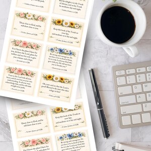 May include: Printed cards with floral borders and Bible verses. The cards feature various flower designs, including roses, sunflowers, and blue flowers. A cup of coffee, a pen, a keyboard, and a notepad are also visible.