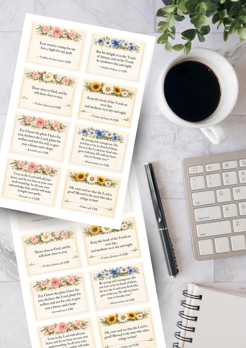 May include: Printed cards with floral borders and Bible verses. The cards feature various flower designs, including roses, sunflowers, and blue flowers. A cup of coffee, a pen, a keyboard, and a notepad are also visible.