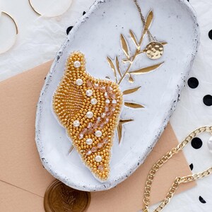 May include: An assortment of jewelry and accessories, including a gold beaded seashell brooch, gold chain necklace, earrings, and bracelet. The items are arranged on a white ceramic dish and a beige envelope with a wax seal. A gold heart is drawn on a white card.