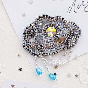 May include: A beaded cloud-shaped brooch with a large yellow center stone and dangling blue beads. The brooch is embellished with various beads and crystals. The background includes cards with handwritten text and star-shaped confetti.