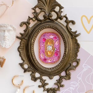 May include: An ornate, antique-style brass frame surrounds a beaded pink and gold brooch. The image also features gold-rimmed glasses, a small bust, gold hoop earrings with white pearl drops, a perfume bottle, and a small jar of gold glitter.