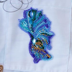 Op de afbeelding: Handgemaakte geborduurde broche in blauw, turquoise en goud, in de vorm van een veer of blad. De broche is versierd met iriserende kralen en heldere stenen. Geplaatst op een witte overhemdzak.