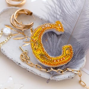 May include: A collection of gold-toned jewelry and accessories. Includes hoop earrings, a beaded letter "C" in yellow and orange, a gold chain necklace, and a decorative feather. Silver star-shaped confetti is scattered around the items.