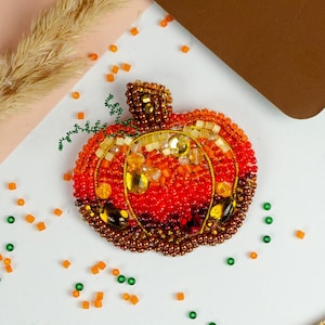 以下が含まれることがあります： オレンジ、赤、茶色のビーズで丁寧に作られた、手作りのカボチャのブローチ。秋をテーマにしたアクセサリーは、茎と緑と金のアクセントを備えた詳細なデザインが特徴です。お祝いの装飾品です。