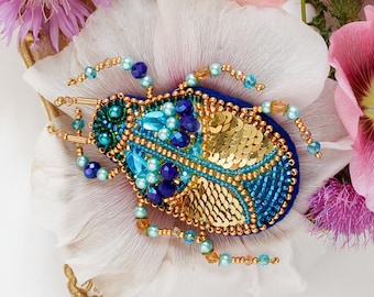 May include: A decorative brooch in the shape of a beetle, featuring blue, gold, and turquoise beads and sequins. The brooch is set on a white flower, surrounded by pink and purple flowers, and a gold-colored ornate frame.