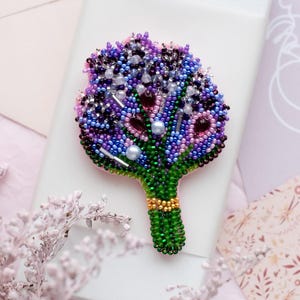 May include: A beaded brooch in the shape of a bouquet of flowers. The brooch features a variety of colors, including purple, blue, green, and pink. The stem is green with a gold band. The brooch is on a white box.