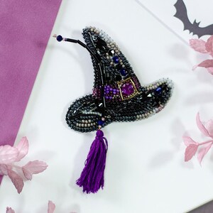 May include: A handcrafted, beaded witch hat brooch in black, purple, and silver. The hat features a purple tassel and a square, purple jewel. The brooch is set against a white background with decorative elements, including paper bats and pink leaves.