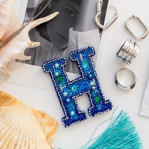 May include: An assortment of jewelry and accessories, including a beaded blue letter "H" brooch, silver necklaces, earrings, and rings. A turquoise tassel and seashells are also present, creating a beachy theme.