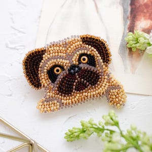 Può includere: Spilla a forma di carlino realizzata a mano con perline marroni, beige e nere. Il carlino è sdraiato, con grandi occhi rotondi e una dettagliata trama di perline. Sullo sfondo un'illustrazione ad acquerello di cani.