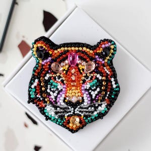 May include: A beaded tiger head brooch in a white box. The brooch features a black outline with orange, red, yellow, green, and purple beads. The tiger's face is detailed with various bead colors and textures.
