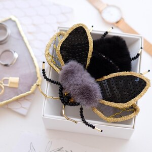 May include: A decorative insect brooch with black sequined wings edged in gold, a grey fur body, and beaded antennae. The brooch is presented in a white box. The design suggests a luxury item.