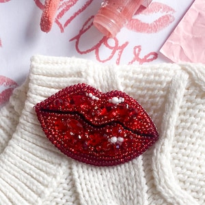 May include: A cream cable-knit jumper featuring a red beaded lips brooch. Red beads are scattered on the jumper. Lip gloss and lipstick kiss prints on paper create a romantic theme. A beauty and fashion-themed image.