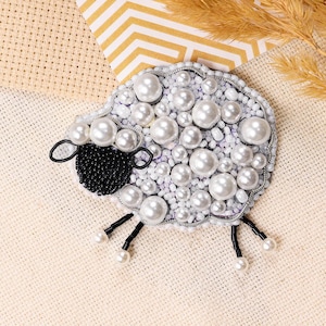 Puede incluir: Broche con forma de oveja con cuerpo de cuentas blancas y grandes detalles de perlas. La oveja tiene una cara de cuentas negras y patas negras con pies de perlas. El broche está sobre un fondo neutro con trigo decorativo.