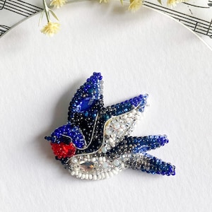 May include: A beaded bird brooch in shades of blue, white, and red, with a detailed design. The bird is set on a white circular surface, with sheet music and floral accents in the background. The brooch is a decorative accessory.