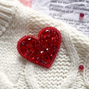 以下が含まれることがあります： クリーム色のケーブル編みセーターのクローズアップ。キラキラ光る赤いハート型のブローチが飾られています。小さな赤いビーズが散りばめられています。背景にはテキストが書かれたヴィンテージペーパーがあります。
