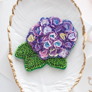 Puede incluir: Broche artesanal con forma de flor de hortensia. El broche presenta lentejuelas moradas y rosas, detalles azules y hojas verdes con cuentas. Descansa sobre una fuente blanca en forma de pluma con borde dorado.
