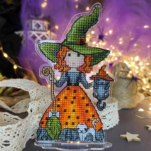 May include: A cross-stitched Halloween decoration of a witch in a green hat and orange dress, holding a staff and lantern. A white cat sits at her feet. The background features fairy lights and a purple backdrop.