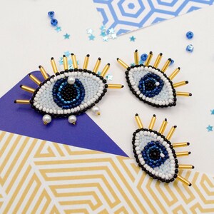 May include: Three beaded eye-shaped ornaments with blue, white, and gold accents. The eyes have a dark blue pupil and are surrounded by gold-colored spikes. Small blue beads and star-shaped confetti are scattered around the ornaments.