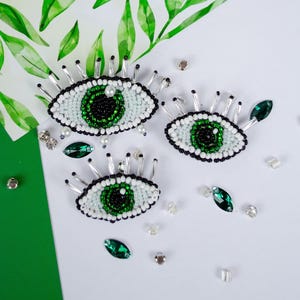 Peut inclure: Trois broches en forme d'œil perlées, avec des centres verts et noirs, entourées de perles blanches. Les broches ont des accents argentés et sont parsemées de gemmes vertes et transparentes. Le fond est blanc et vert avec un motif de feuilles à l'aquarelle.